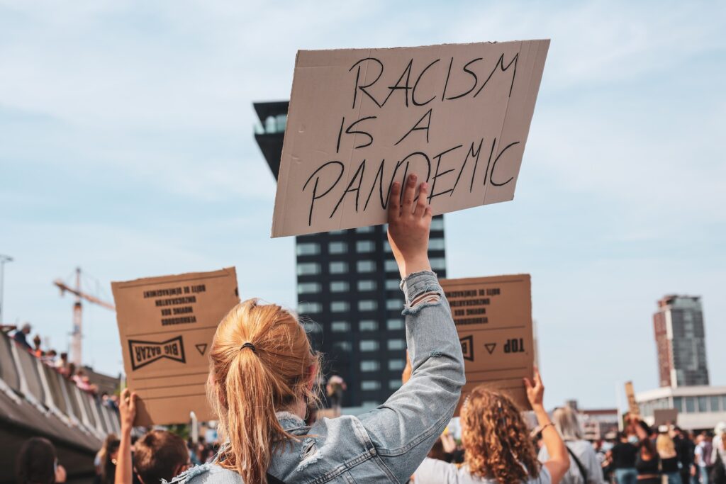 Plakat głoszący, że rasizm jest pandemią trzymany w dłoni przez jasnowłosą dziewczynę odwróconą plecami do obiektywu w trakcie demonstracji.
