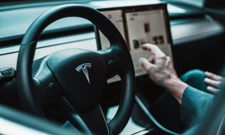 Wnętrze samochodu Tesla. Kierownica z logo firmy i dłoń mężczyzny skierowana na komputer pokładowy.
